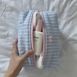 Puede incluir: Un neceser a rayas azules y blancas con cremallera rosa, que contiene un frasco blanco de producto capilar "gisou" y un paño blanco. Un pequeño lazo rosa está sujeto al lateral de la bolsa.