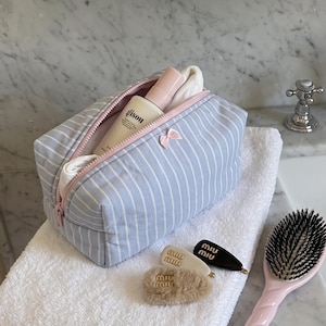 May include: A light blue and white striped fabric wash bag with a pink zip and a small pink bow detail. The bag contains a Gisou bottle and white fabric. Nearby are two Miu Miu hair clips, a fuzzy hair accessory, and a pink hairbrush.