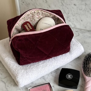 May include: A quilted burgundy velvet cosmetic bag with a light pink zipper and floral interior lining, containing a white egg-shaped hand cream and a red lipstick. Nearby are a pink hairbrush, a Dior blush, and two Chanel compacts.