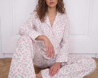 May include: A two-piece pyjama set featuring a long-sleeved button-down shirt with a notched collar and matching wide-leg trousers. The fabric is white with an all-over floral print consisting of small pink roses and green leaves with pink piping.