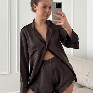 May include: A dark brown satin pajama set. The set includes a long-sleeved button-down shirt with a chest pocket and matching shorts with a ruffled hem. The model is taking a selfie.