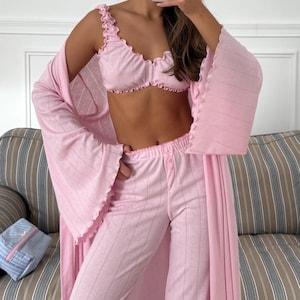 May include: A light pink pajama set with a matching robe. The set includes a ruffled crop top, pajama pants, and a long-sleeved robe. The fabric has a subtle dotted pattern. The robe has wide sleeves.