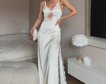 May include: A floor-length white satin slip dress featuring thin spaghetti straps, a cutout bodice with white lace trim, and a high side slit accented with matching lace along the edge. The dress has a smooth, lustrous finish and a fluid silhouette.