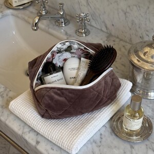 May include: A brown quilted velvet cosmetic bag with a white zipper and a floral interior lining. The bag is filled with a Byredo Mojave Ghost perfume bottle, a white Chanel La Cr&egrave;me Main hand cream, a small tube, and a dark brown hairbrush.