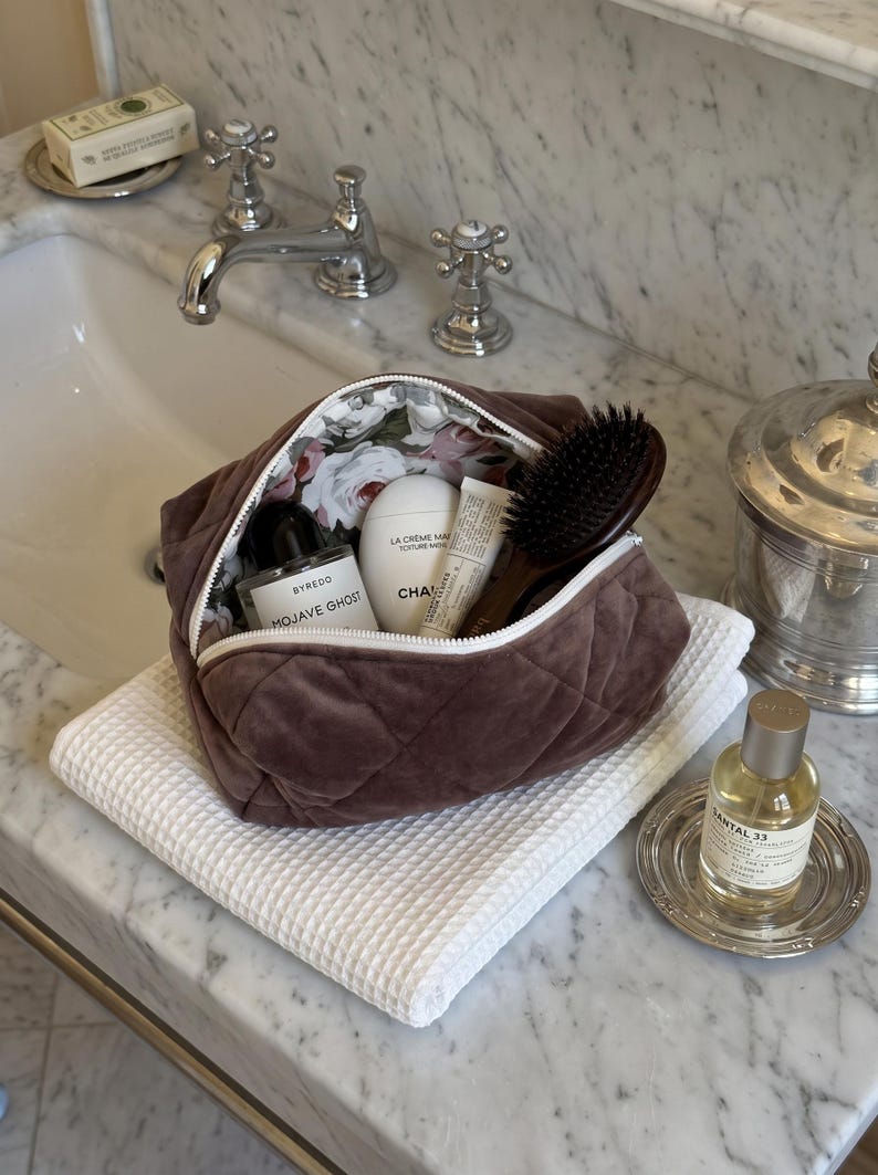 May include: A brown quilted velvet cosmetic bag with a white zipper and a floral interior lining. The bag is filled with a Byredo Mojave Ghost perfume bottle, a white Chanel La Cr&egrave;me Main hand cream, a small tube, and a dark brown hairbrush.