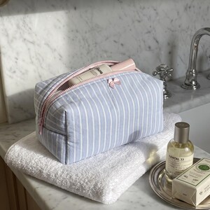 May include: A light blue and white striped rectangular toiletry bag with a pink zip closure and a small pink bow detail on the front. The bag is partially open, revealing a white bottle inside, and it rests on a folded white towel.