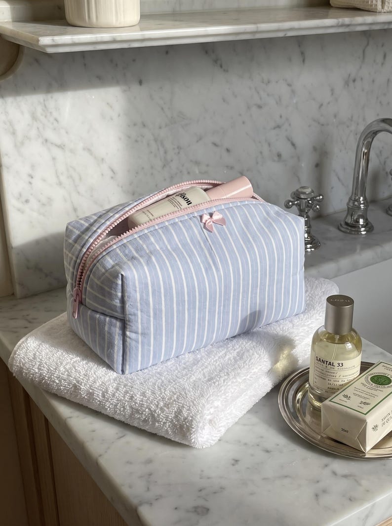 May include: A light blue and white striped rectangular toiletry bag with a pink zip closure and a small pink bow detail on the front. The bag is partially open, revealing a white bottle inside, and it rests on a folded white towel.