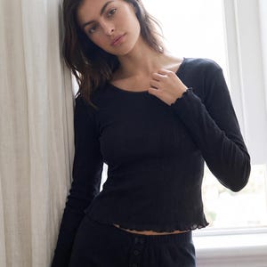 May include: A black long-sleeved top featuring a round neckline with delicate trim, a subtle vertical ribbed texture, and lettuce-edge detailing along the cuffs and hemline. The garment is paired with matching black bottoms that have a button-front placket.
