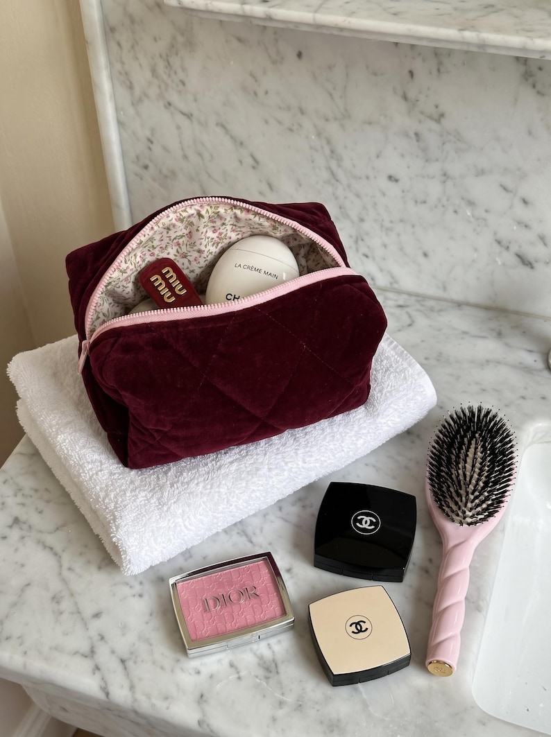 May include: A quilted burgundy velvet cosmetic bag with a light pink zipper and floral interior lining, containing a white egg-shaped hand cream and a red lipstick. Nearby are a pink hairbrush, a Dior blush, and two Chanel compacts.