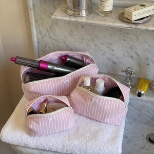 May include: A set of three light pink and white striped fabric pouches with white zips and small white bows. The largest pouch contains a grey and fuchsia hair styling tool, the medium pouch holds bottles and a brush, and the smallest holds lipstick.