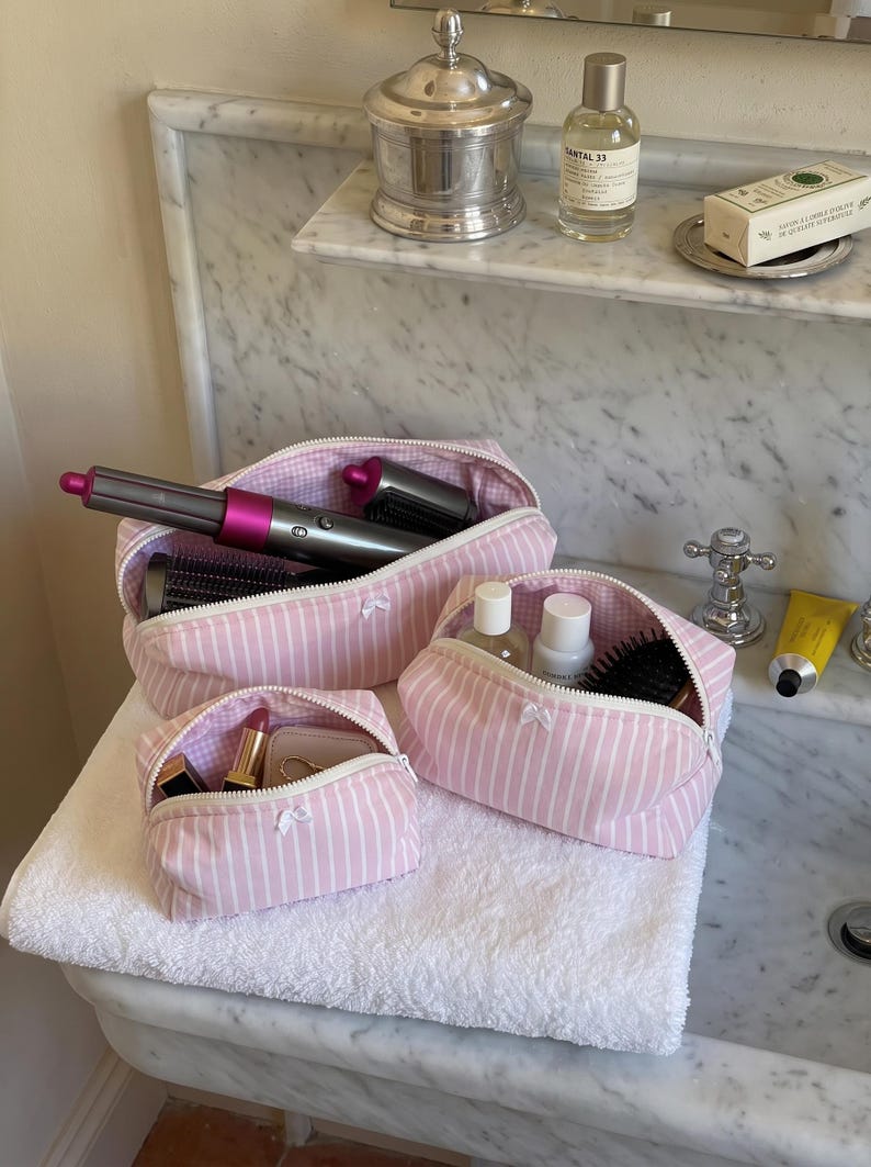 May include: A set of three light pink and white striped fabric pouches with white zips and small white bows. The largest pouch contains a grey and fuchsia hair styling tool, the medium pouch holds bottles and a brush, and the smallest holds lipstick.