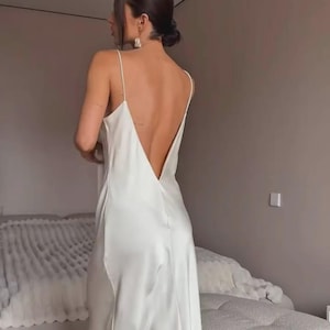 May include: A white satin slip dress with a low back and spaghetti straps. The dress is floor-length and has a simple, elegant design. The model is wearing the dress in a bedroom setting.