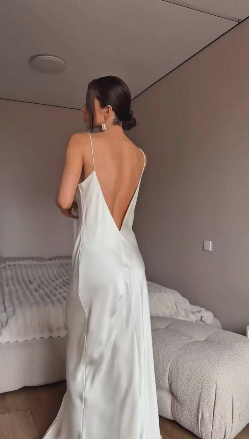 May include: A white satin slip dress with a low back and spaghetti straps. The dress is floor-length and has a simple, elegant design. The model is wearing the dress in a bedroom setting.