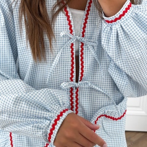 Puede incluir: Bata con estampado de cuadros vichy azul claro y blanco con ribete rojo. La bata presenta un ribete rojo festoneado a lo largo de los bordes, pu&ntilde;os y cierres de lazo. Las mangas son largas y fruncidas en los pu&ntilde;os.