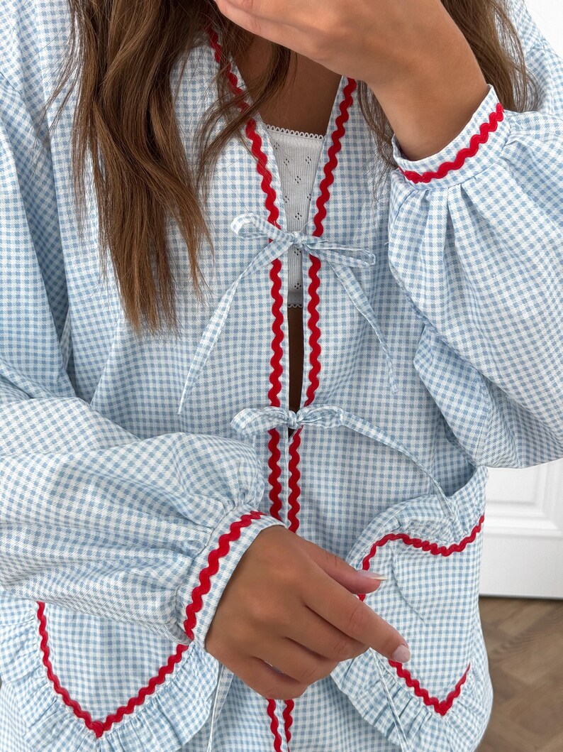 Puede incluir: Bata con estampado de cuadros vichy azul claro y blanco con ribete rojo. La bata presenta un ribete rojo festoneado a lo largo de los bordes, pu&ntilde;os y cierres de lazo. Las mangas son largas y fruncidas en los pu&ntilde;os.