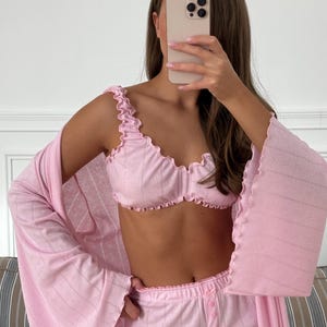 May include: A light pink pajama set featuring a ruffled bralette, matching pants, and a long robe. The set has a subtle pattern and is made of a soft, comfortable material. The pajama set is designed for lounging and relaxation.