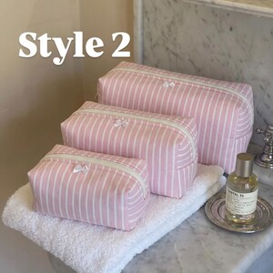 May include: Three rectangular cosmetic bags in varying sizes, featuring a light pink and white vertical striped pattern. Each bag has a white zip closure and a small white bow detail. The text Style 2 is visible in the upper left corner.