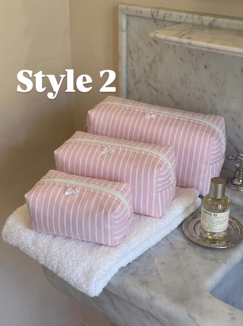May include: Three rectangular cosmetic bags in varying sizes, featuring a light pink and white vertical striped pattern. Each bag has a white zip closure and a small white bow detail. The text Style 2 is visible in the upper left corner.