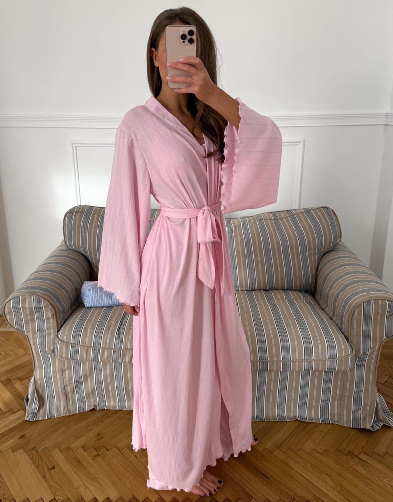 May include: A light pink robe with a matching belt. The robe has wide sleeves with a ruffled edge and a wrap-around design. The model is wearing the robe in front of a striped couch.