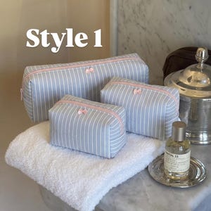 May include: Three light blue and white striped cosmetic bags of varying sizes, each featuring a pale pink zip closure and a small pink bow detail. The text "Style 1" is visible in the upper left corner of the image.