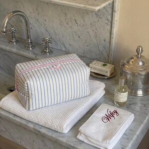 May include: A rectangular toiletry bag with blue and white vertical stripes and a pink zip closure. The bag rests on a white waffle-weave towel next to a smaller white hand towel embroidered with the word Wifey in dark red script lettering.