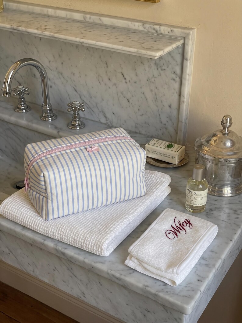 May include: A rectangular toiletry bag with blue and white vertical stripes and a pink zip closure. The bag rests on a white waffle-weave towel next to a smaller white hand towel embroidered with the word Wifey in dark red script lettering.