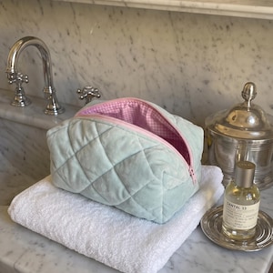 May include: A quilted mint green velvet cosmetic bag with a zipper closure. The interior is lined with a pink and white gingham fabric. The bag has a soft, plush texture and a structured rectangular shape with rounded corners and a top opening.