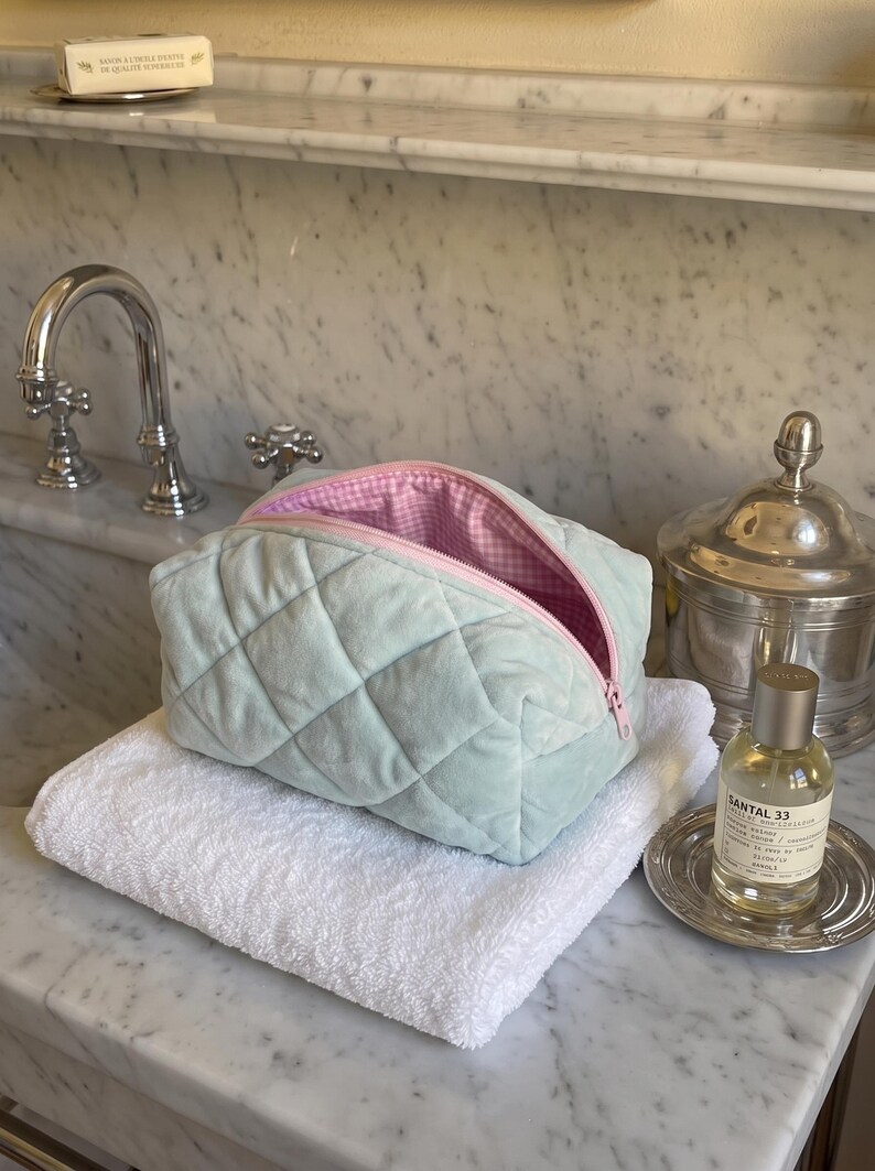 May include: A quilted mint green velvet cosmetic bag with a zipper closure. The interior is lined with a pink and white gingham fabric. The bag has a soft, plush texture and a structured rectangular shape with rounded corners and a top opening.