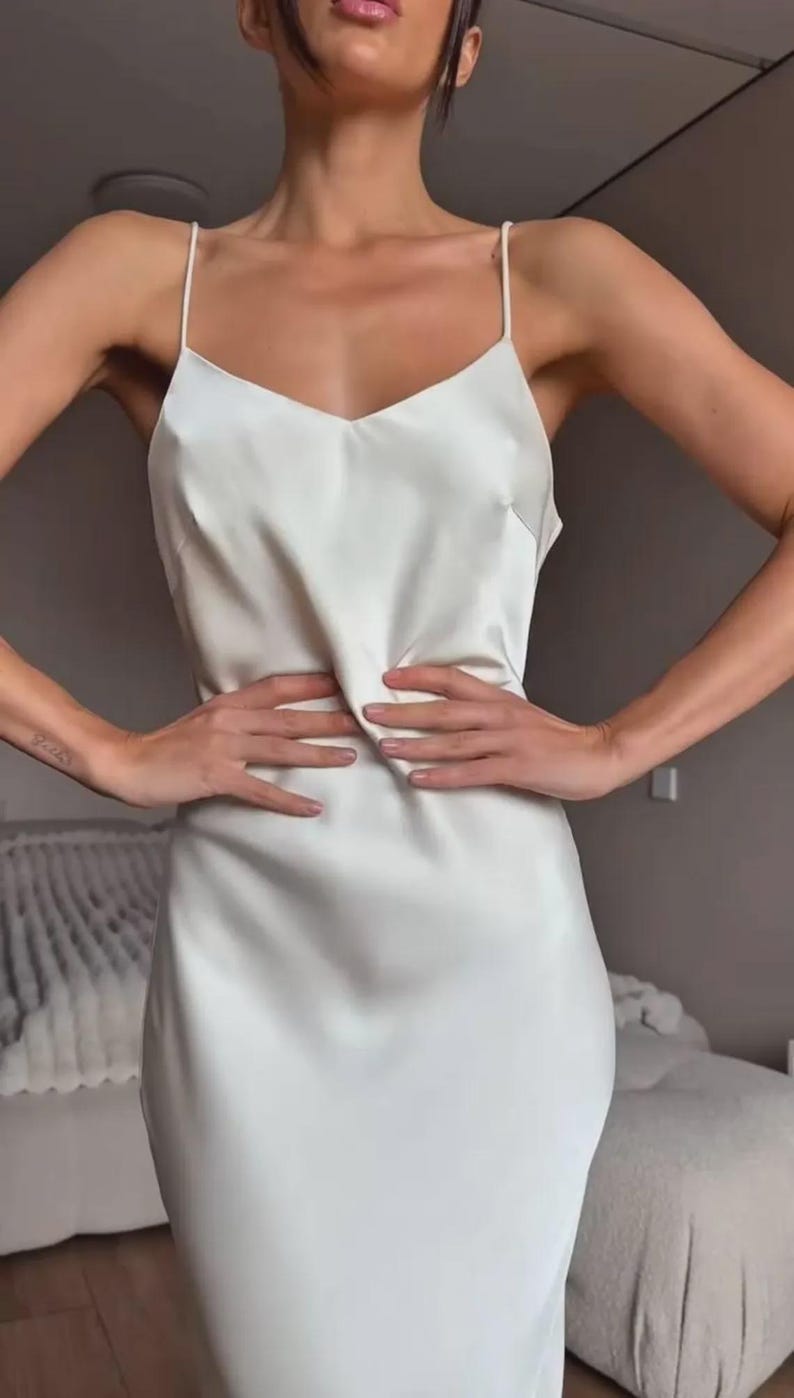 May include: A white satin slip dress with spaghetti straps and a V-neck. The dress is knee-length and has a simple, elegant design. The fabric appears smooth and drapes softly. The dress is a solid color and has a minimalist aesthetic.