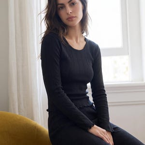 May include: A black long-sleeve top featuring a ribbed texture, a round neckline with delicate trim, and lettuce-edge detailing along the cuffs and hem. The garment is constructed from a lightweight, fitted fabric that contours to the upper body.