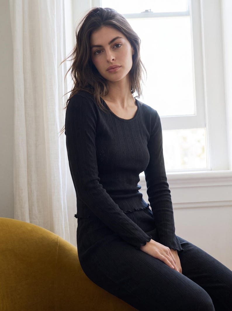 May include: A black long-sleeve top featuring a ribbed texture, a round neckline with delicate trim, and lettuce-edge detailing along the cuffs and hem. The garment is constructed from a lightweight, fitted fabric that contours to the upper body.