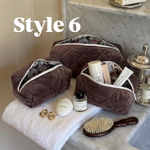 May include: Three dark brown quilted velvet cosmetic bags with white zippers and floral interior lining. The bags are arranged with various personal care items including a Chanel hand cream, Byredo perfume, and a wooden hair brush nearby.