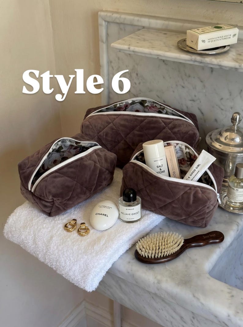 May include: Three dark brown quilted velvet cosmetic bags with white zippers and floral interior lining. The bags are arranged with various personal care items including a Chanel hand cream, Byredo perfume, and a wooden hair brush nearby.