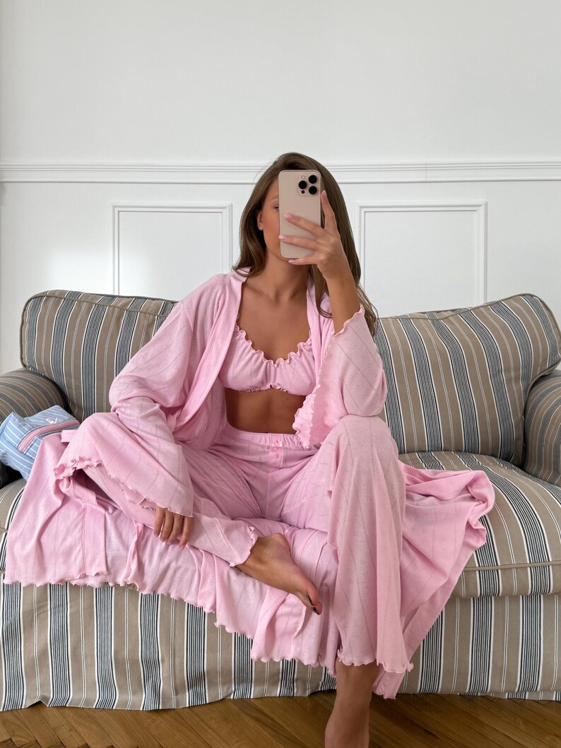 May include: A light pink pajama set consisting of a ruffled-edge robe, a bralette, and wide-leg pants. The set is made of a soft, flowing fabric. The model is sitting on a striped sofa.