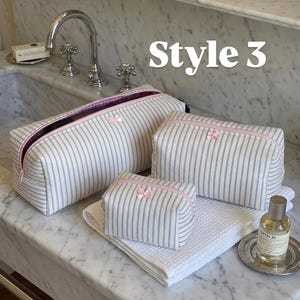May include: Three rectangular toiletry bags in varying sizes, featuring a blue and white vertical striped pattern, pink zipper closures, and a small pink bow detail on the front of each. The text Style 3 is visible in the upper right area.