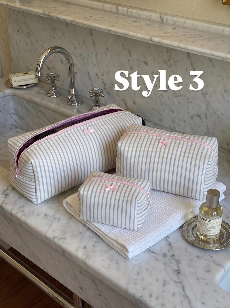 May include: Three rectangular toiletry bags in varying sizes, featuring a blue and white vertical striped pattern, pink zipper closures, and a small pink bow detail on the front of each. The text Style 3 is visible in the upper right area.
