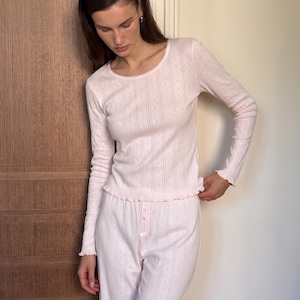 May include: A light pink long-sleeve top and matching pants set. The top features a round neckline, long sleeves with lettuce-edge cuffs, and a vertical pointelle knit pattern. The pants have a button-front placket and a matching vertical pointelle knit.