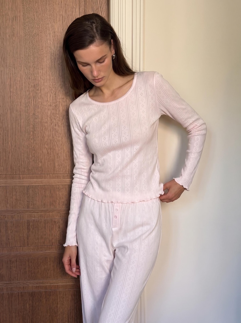 May include: A light pink long-sleeve top and matching pants set. The top features a round neckline, long sleeves with lettuce-edge cuffs, and a vertical pointelle knit pattern. The pants have a button-front placket and a matching vertical pointelle knit.