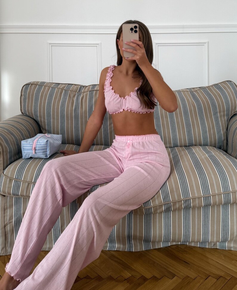 May include: Pink pajama set with a ruffled crop top and matching pants. The set features a striped pattern and is made of a soft fabric. A blue and white striped cosmetic bag is on the sofa.