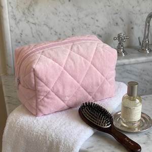 May include: A light pink quilted cosmetic bag with a zipper closure, a dark brown wooden hairbrush with black bristles, and a glass bottle of Santal 33 perfume with a silver cap, all resting on a white textured towel.
