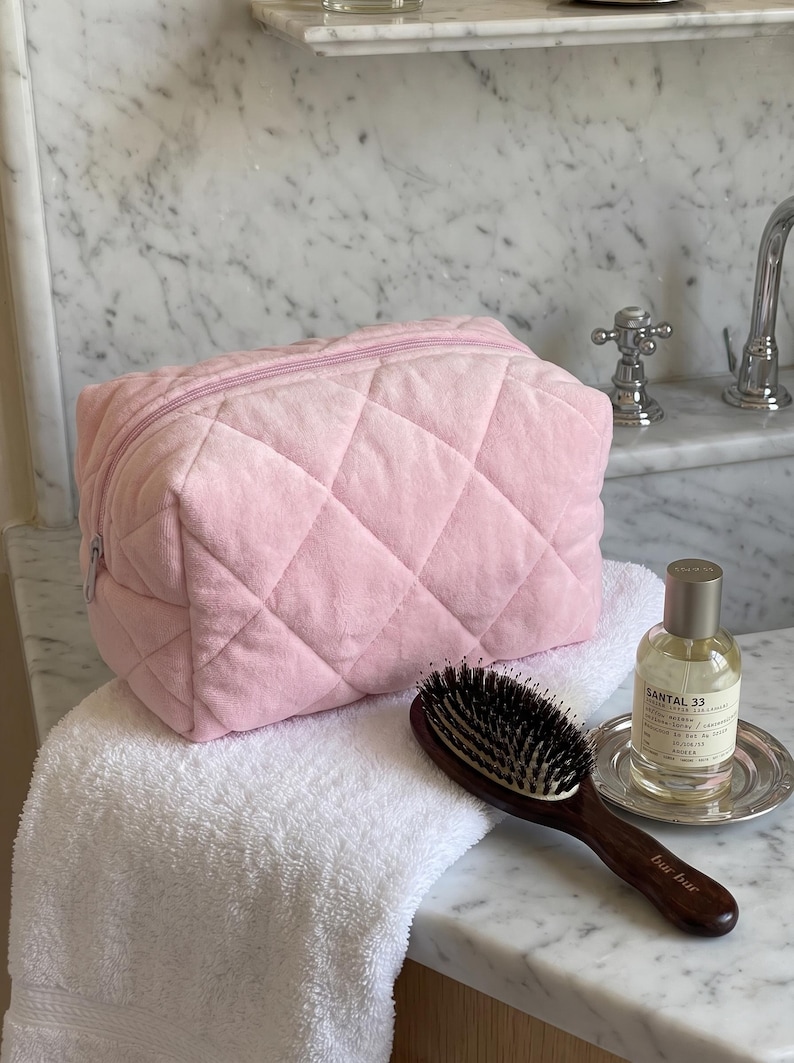 May include: A light pink quilted cosmetic bag with a zipper closure, a dark brown wooden hairbrush with black bristles, and a glass bottle of Santal 33 perfume with a silver cap, all resting on a white textured towel.