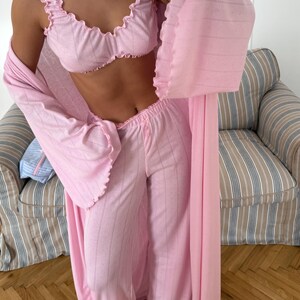 May include: A light pink pajama set with a matching robe. The set includes a ruffled-edge bra top, wide-leg pants, and a long robe. The fabric has a subtle striped pattern. The robe has wide sleeves and a delicate, open weave design.