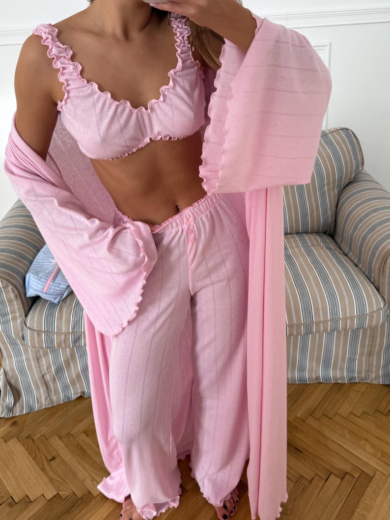 May include: A light pink pajama set with a matching robe. The set includes a ruffled-edge bra top, wide-leg pants, and a long robe. The fabric has a subtle striped pattern. The robe has wide sleeves and a delicate, open weave design.