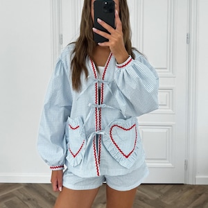 Puede incluir: Conjunto de pijama de cuadros vichy azul claro y blanco. La parte superior tiene cuello en V con ribete rojo y bolsillos en forma de coraz&oacute;n con ribete rojo. Los pantalones cortos coinciden con el patr&oacute;n y el ribete de la parte superior. Las mangas tienen pu&ntilde;os el&aacute;sticos con ribete rojo.