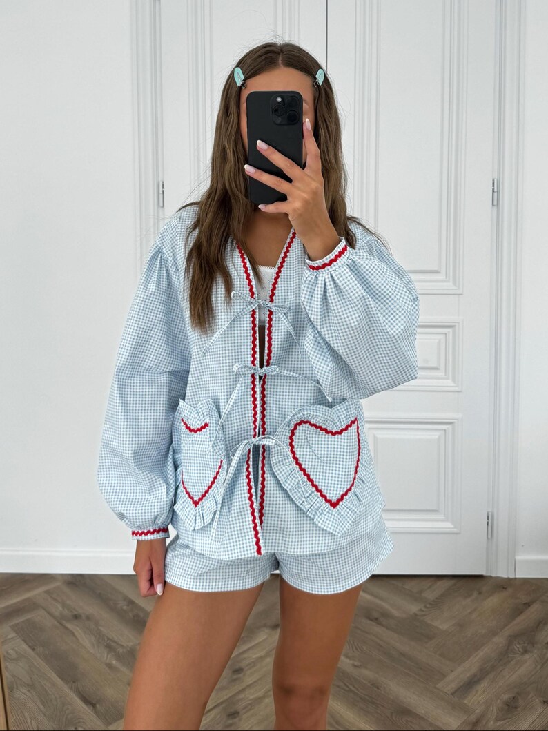 Puede incluir: Conjunto de pijama de cuadros vichy azul claro y blanco. La parte superior tiene cuello en V con ribete rojo y bolsillos en forma de coraz&oacute;n con ribete rojo. Los pantalones cortos coinciden con el patr&oacute;n y el ribete de la parte superior. Las mangas tienen pu&ntilde;os el&aacute;sticos con ribete rojo.