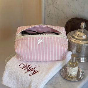 May include: A pink and white striped fabric pouch with a white zip and a small white bow on the front. The pouch is partially open, revealing black spectacles inside. The pouch sits on a white towel embroidered with the word Wifey in red.