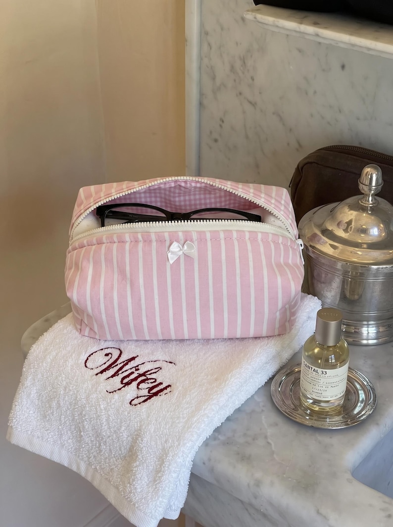 May include: A pink and white striped fabric pouch with a white zip and a small white bow on the front. The pouch is partially open, revealing black spectacles inside. The pouch sits on a white towel embroidered with the word Wifey in red.
