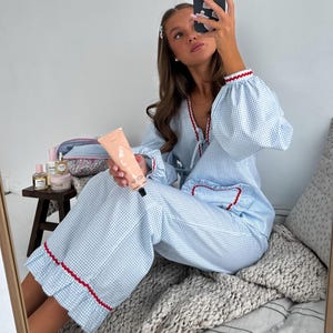 Puede incluir: Conjunto de pijama de cuadros vichy azul claro y blanco con ribete rojo. La parte superior tiene mangas largas con pu&ntilde;os con volantes. Los pantalones tienen pernera ancha y dobladillo con volantes. Una persona sostiene un tubo de loci&oacute;n y un tel&eacute;fono.