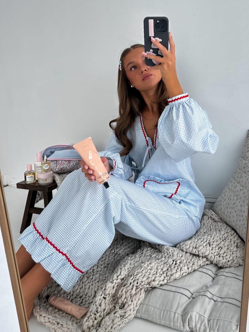 Puede incluir: Conjunto de pijama de cuadros vichy azul claro y blanco con ribete rojo. La parte superior tiene mangas largas con pu&ntilde;os con volantes. Los pantalones tienen pernera ancha y dobladillo con volantes. Una persona sostiene un tubo de loci&oacute;n y un tel&eacute;fono.