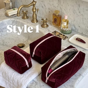 May include: Three burgundy velvet cosmetic bags with a quilted diamond pattern and light pink zippers. The bags vary in size, with one open bag containing a grey hair styling tool. The text "Style 1" is visible in white on the left side.